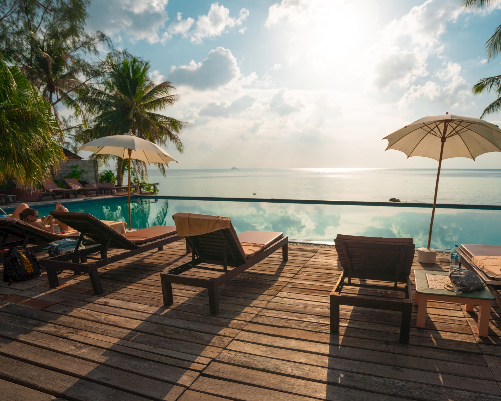 A beachfront resort scene with lounge chairs, a small pool, umbrellas, palm trees, and a calm ocean under bright clouds.