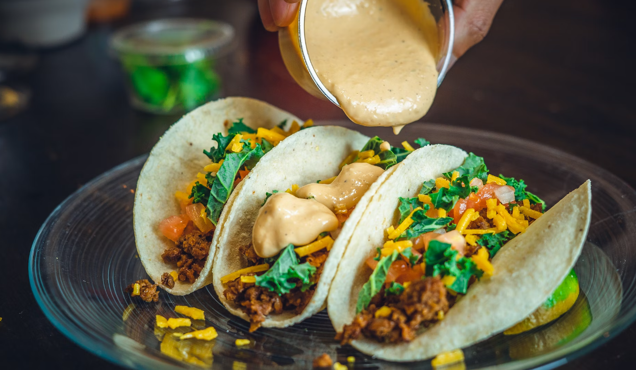 Three tacos on a plate with shredded fillings and cilantro, with creamy sauce being poured over them.
