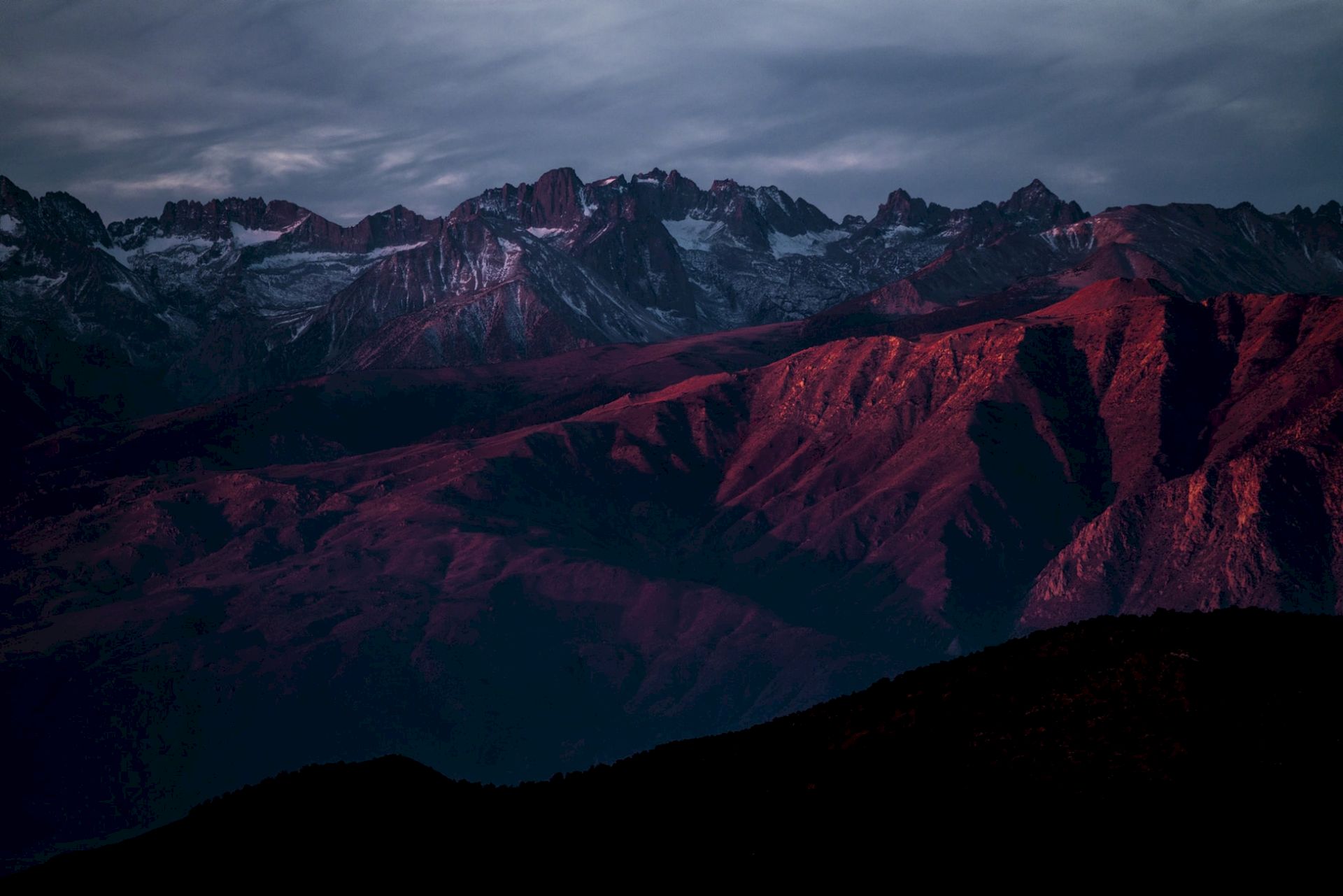 A dramatic mountain range at dusk, snow patches and dark ridges lit by a red sunset glow under stormy clouds.