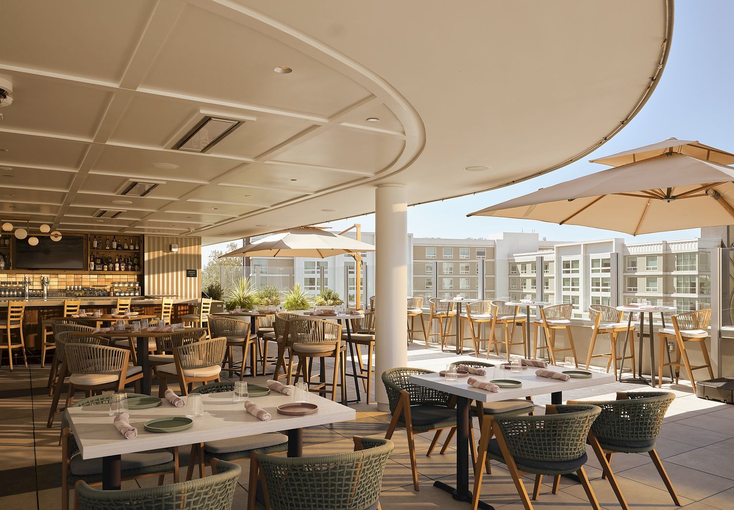 A sunny hotel patio dining area with wooden tables, wicker chairs, umbrellas, and a bar in the background.