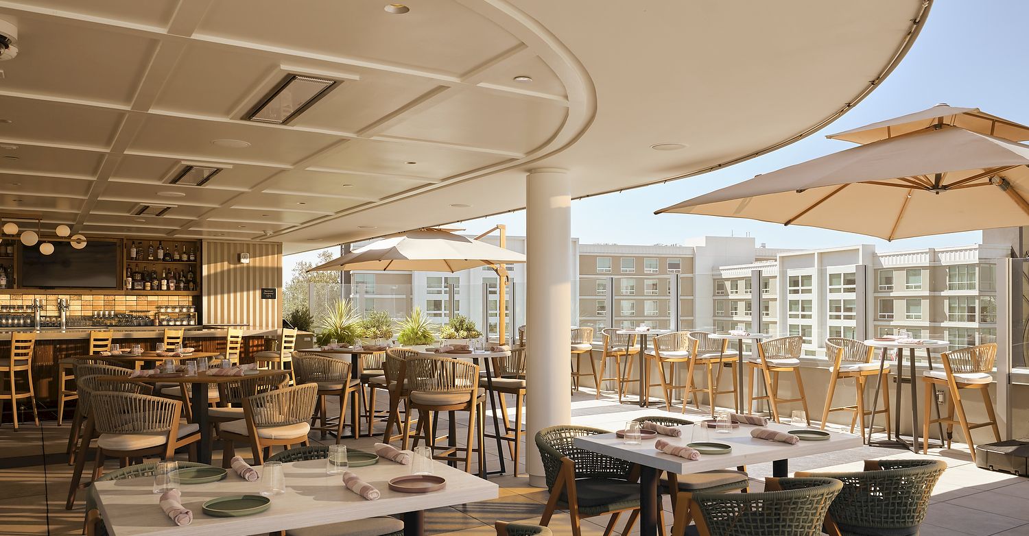 A sunny hotel patio dining area with wooden tables, wicker chairs, umbrellas, and a bar in the background.