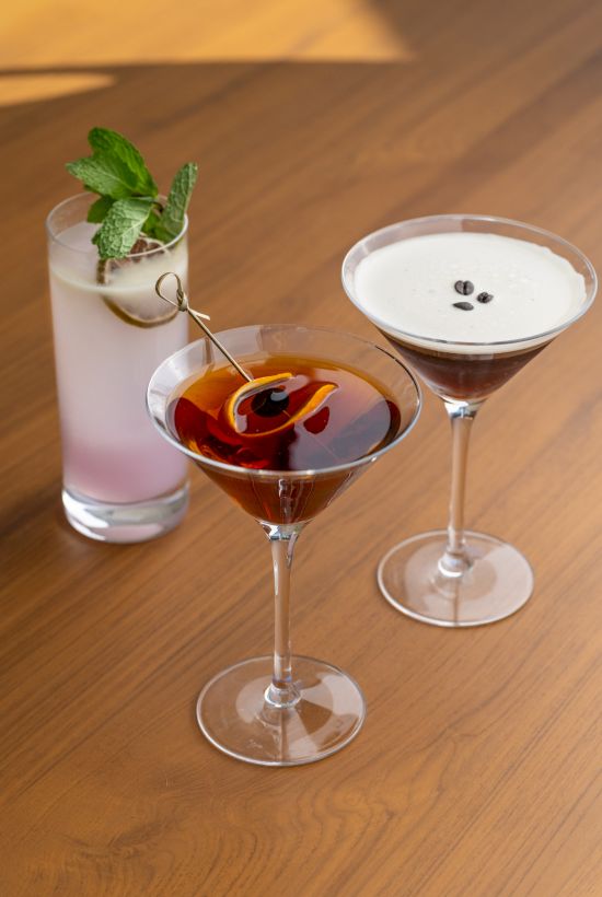 Three cocktails on a wooden bar: a minty tall drink, a cherry-topped coupe, and a creamy martini with coffee beans&mdash;cheerful nightcaps.