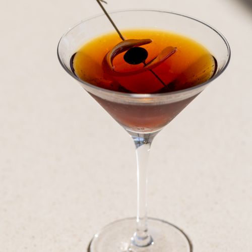 A classic cocktail in a martini glass with an olive stacked on a skewer, sunlight glinting off the rim and long shadow. ending sentence.