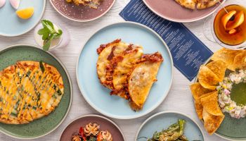 Assorted dishes on colorful plates: slices of quiche, tacos, greens, chips with dip, a burger bun, and plates of roasted veggies for a vibrant brunch spread.