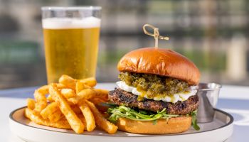 A juicy burger with melted toppings on a bun, a side of crispy fries, and a chilled beer in the background on a plate.