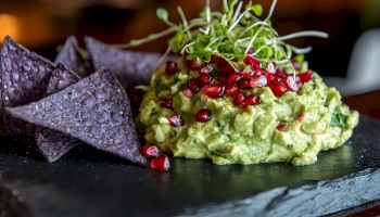 Creamy avocado mash topped with pomegranate seeds and microgreens, served with purple tortilla chips on a black slate plate.