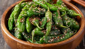 A wooden bowl of sautéed green peppers sprinkled with sesame seeds, likely a Korean-style seasoned peppers. Ends with a period.
