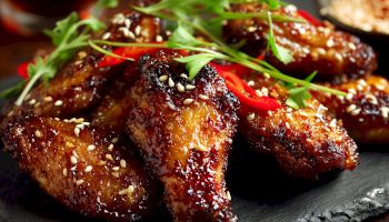 Sticky glazed chicken wings with sesame seeds and greens, glossy caramelized sauce, served on a dark platter.