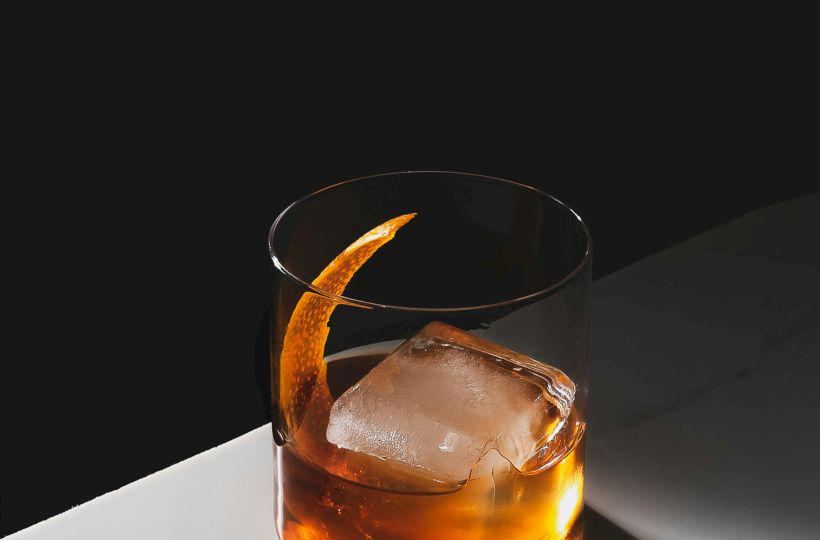 A glass of whiskey with a large ice cube sits on a marble surface, casting a warm glow in a minimalist setting.
