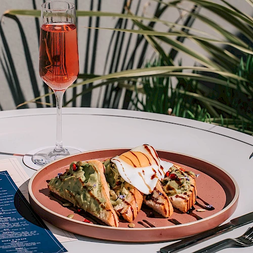 A fancy plate with savory bites, a glass of pink drink, and a blue menu on a white table. Bold flavors ahead.