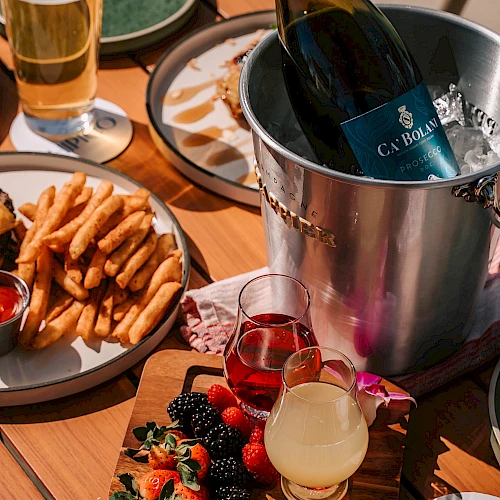 A champagne bottle chilling in a bucket, plus assorted snacks: fries, a veggie-and-fruit board with berries, and two colorful cocktails on a wooden table.