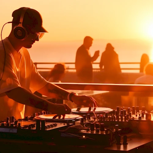 A DJ wearing headphones spins vinyl on a deck at sunset, with silhouettes of people by the railing and a warm orange glow over the scene.