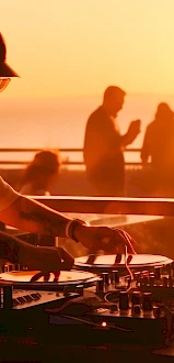 A DJ wearing headphones spins vinyl on a deck at sunset, with people silhouettes enjoying the beachside party in warm orange light.