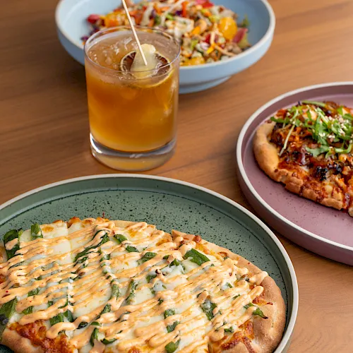 A table of food: pizza slices with creamy drizzle, a side salad, and a glass of orange juice with ice, on plates.