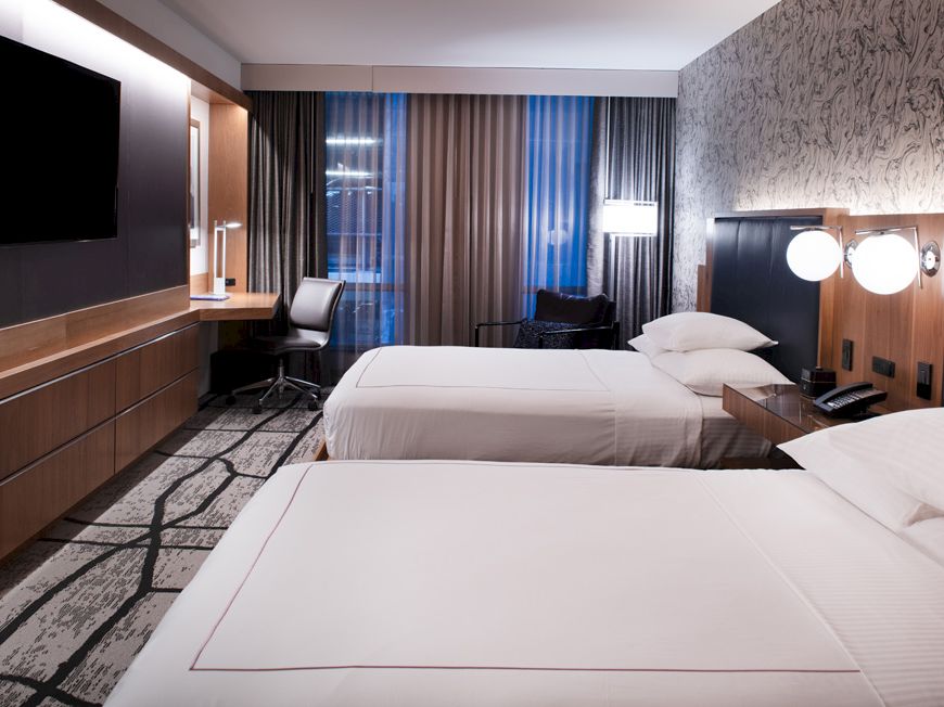 A modern hotel room with two white beds, dark wood panels, a desk and chair, and curtains covering large windows.