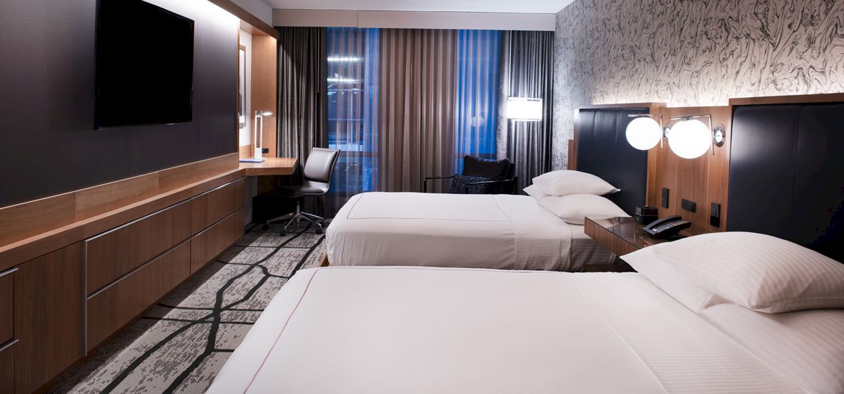 A modern hotel room with two white beds, dark wood panels, a desk and chair, and curtains covering large windows.