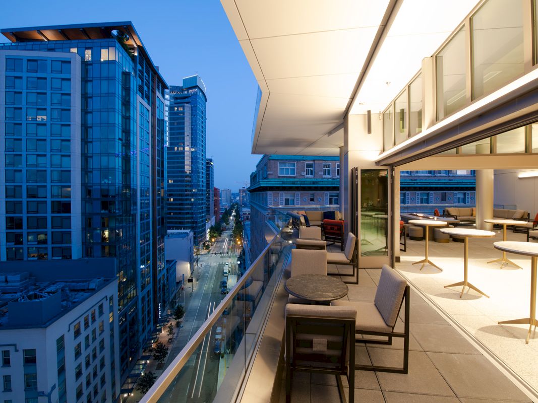 A modern high-rise rooftop terrace with lounge chairs overlooks a busy city street between blue-lit skyscrapers.