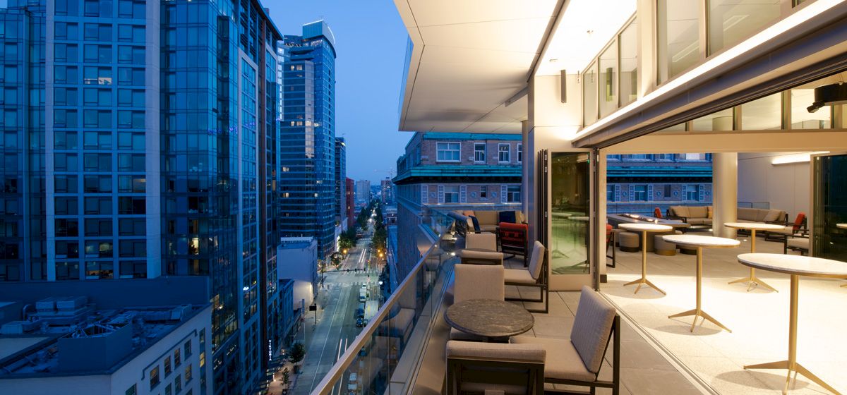 A modern high-rise rooftop terrace with lounge chairs overlooks a busy city street between blue-lit skyscrapers.