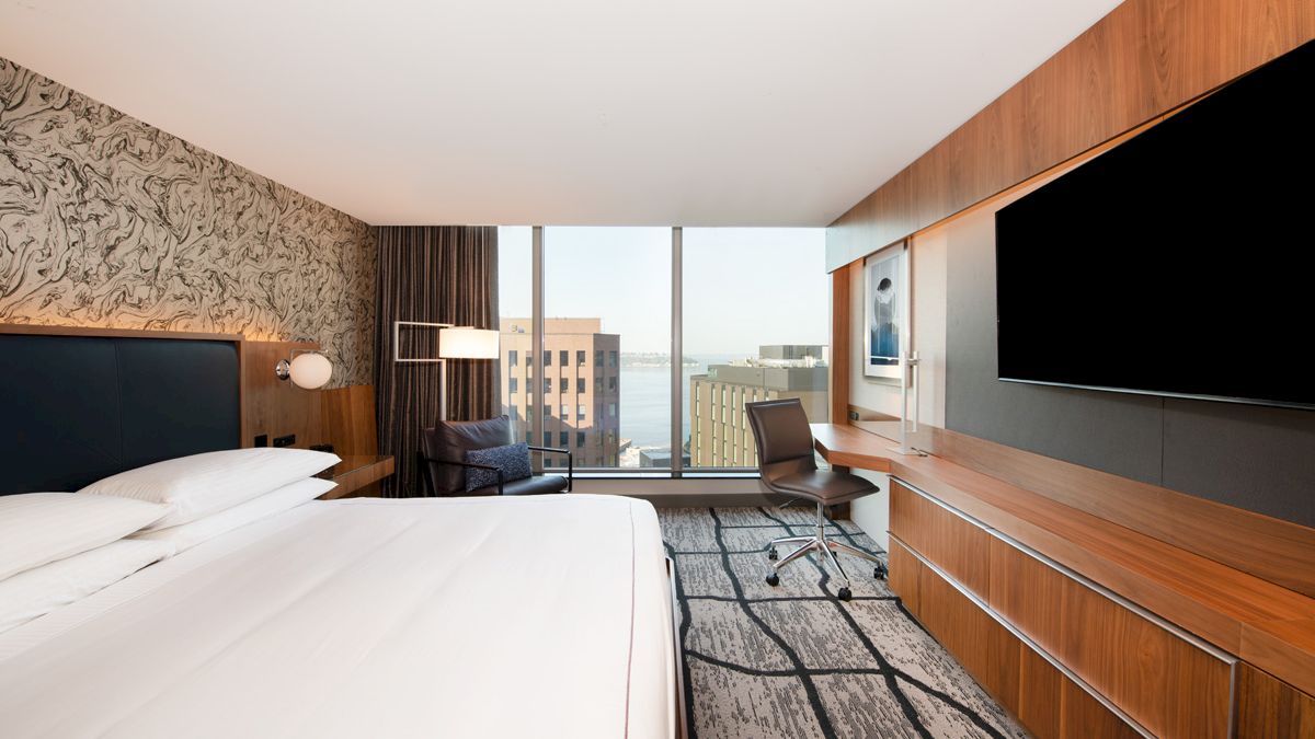 A modern hotel room with a large bed, desk chair, wall-mounted TVs, and floor-to-ceiling windows with a city view.