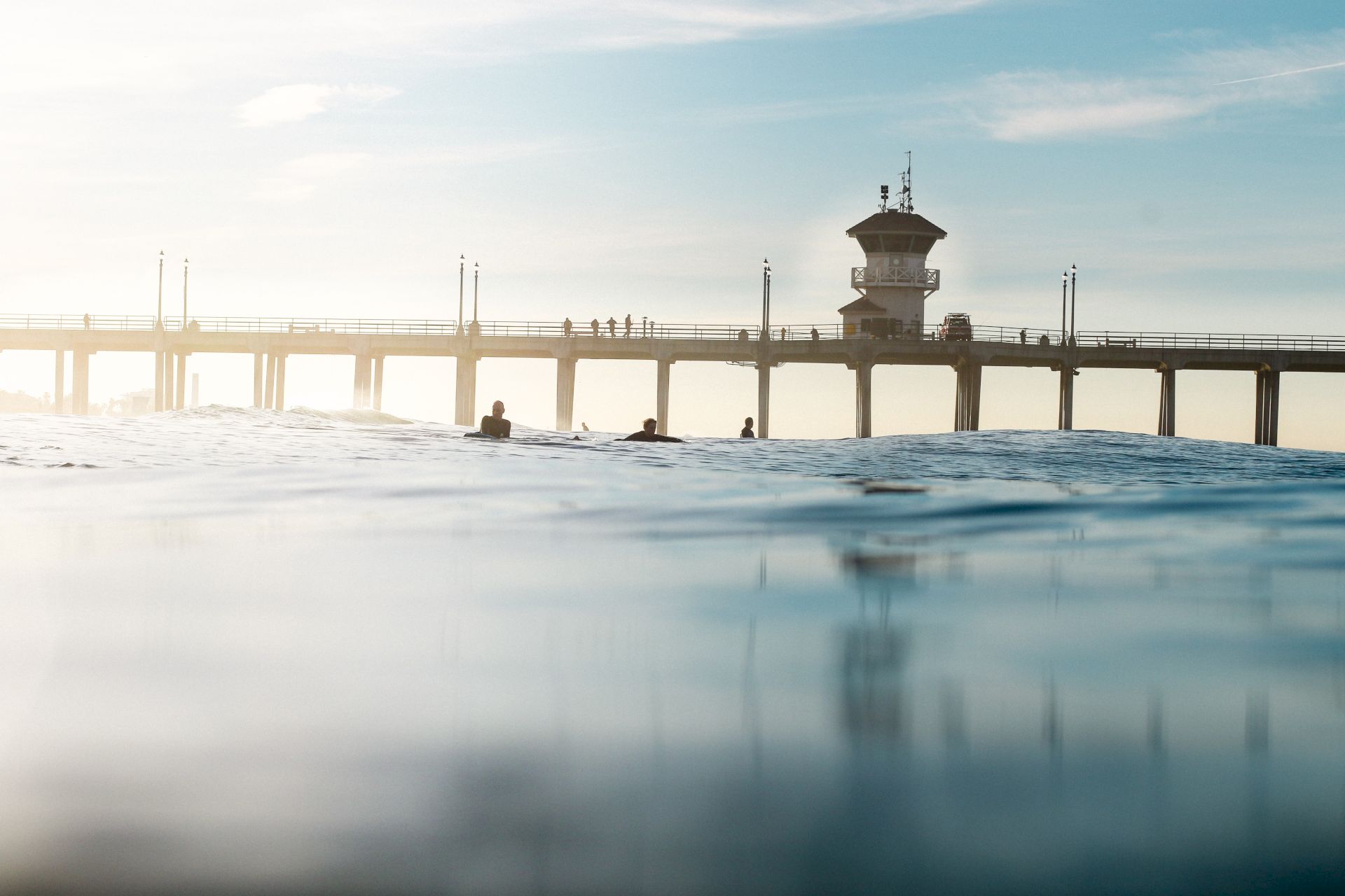 Swimmers float beneath a long pier with a lifeguard tower, under a pale sunset sky over calm water.